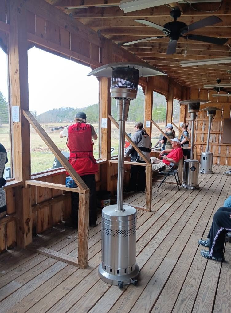 A shooter in a red vest at the rail of a covered shooting pavilion while others wait behind