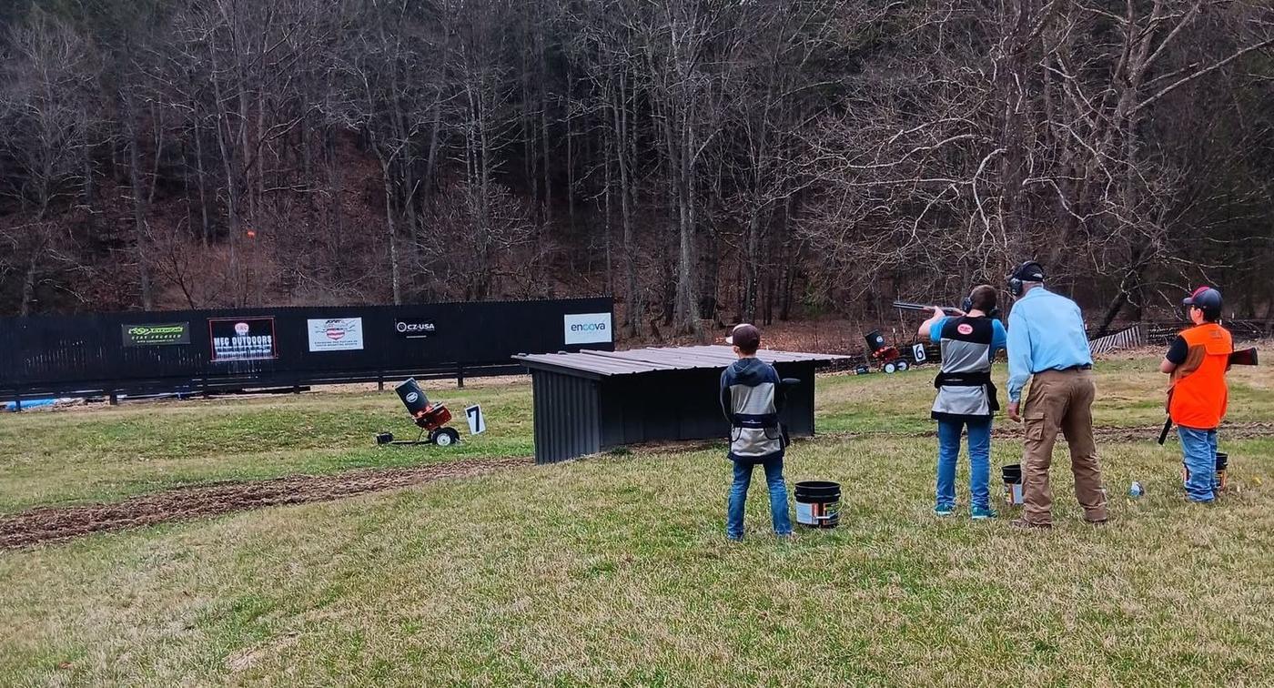 Young shooters waiting their turn at the trap machines while a teammate shoots with an adult standing beside him
